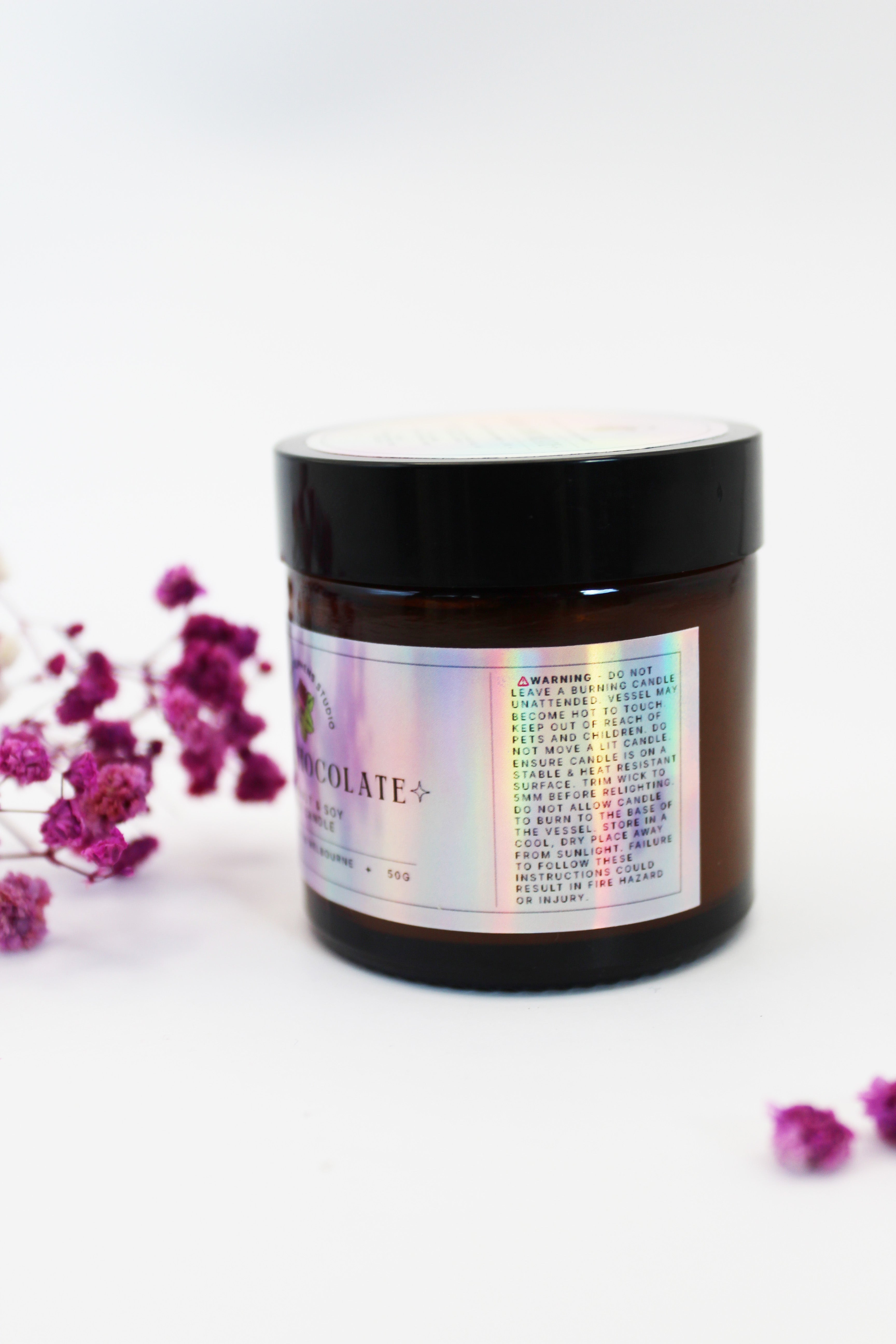 Brown glass candle jar with holographic sticker label, black lid, and pink flowers on a white background