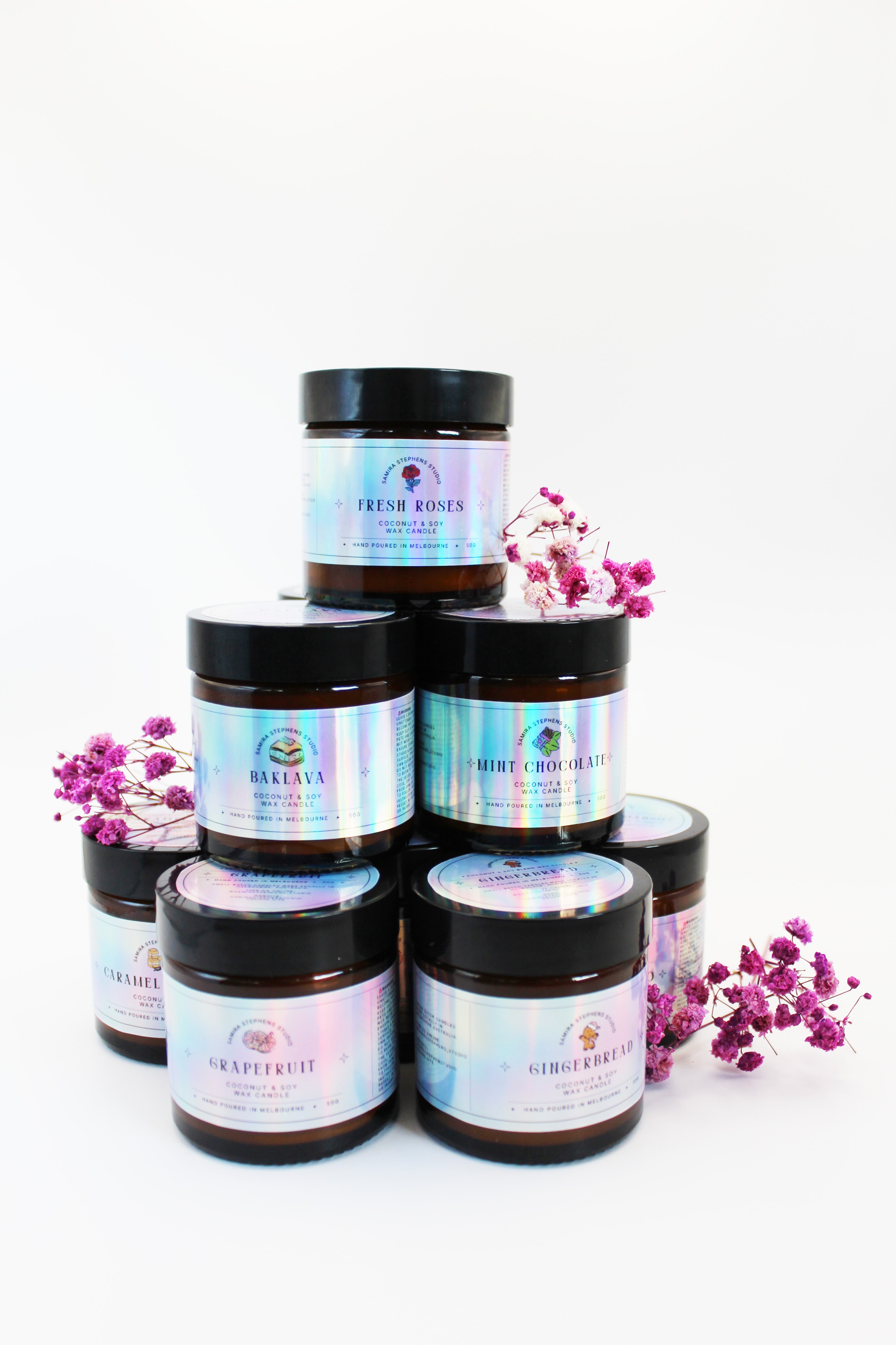Stack of brown candle jars with black lids and colorful labels on a white background with purple flowers